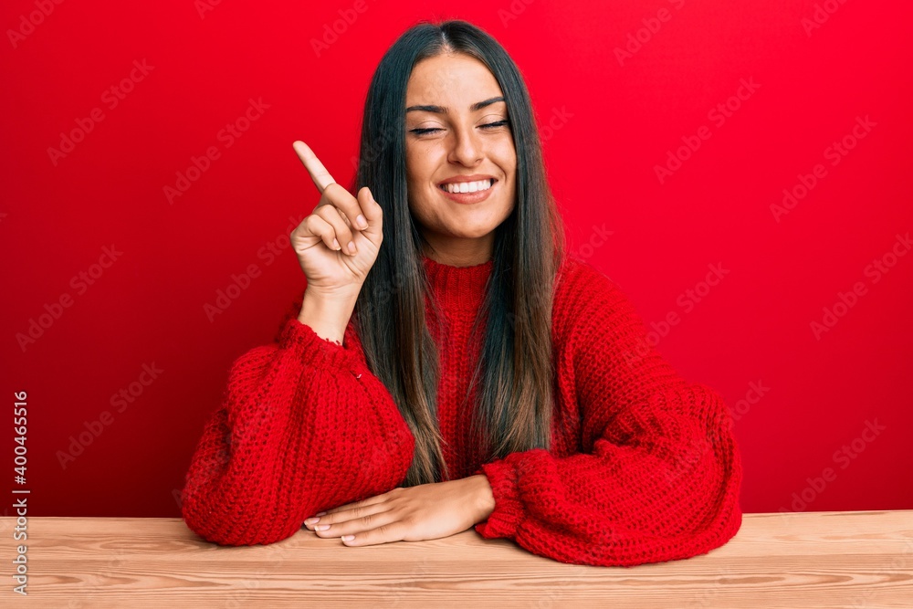 Beautiful hispanic woman wearing casual clothes sitting on the table showing and pointing up with finger number one while smiling confident and happy. Beautiful hispanic woman wearing casual clothes sitting on the table showing and pointing up with finger number one while smiling confident and happy.