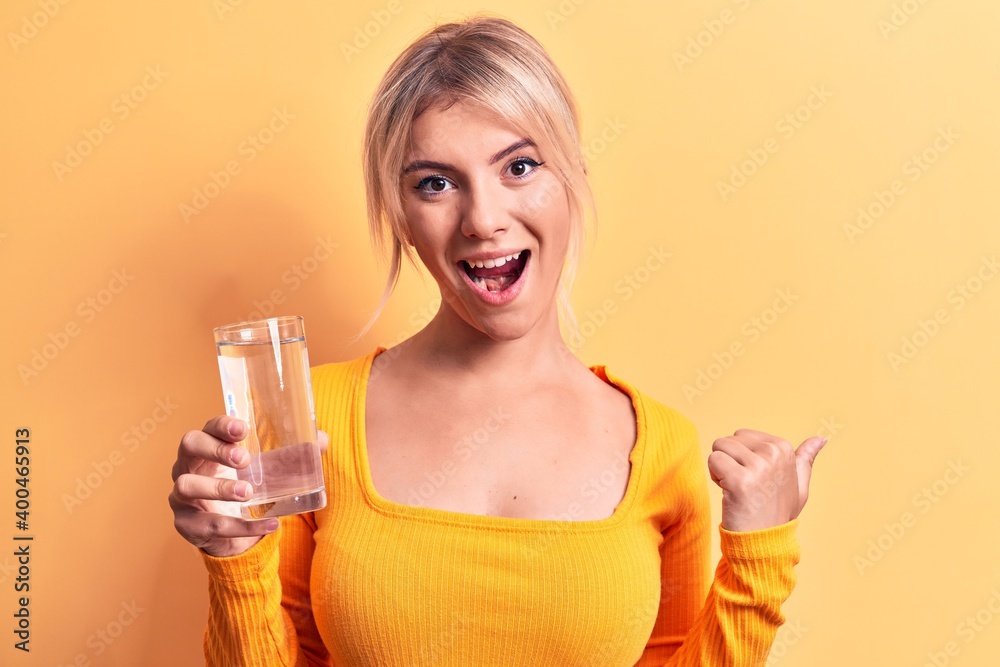 Young beautiful blonde woman drinking glass of water over isolated yellow background pointing thumb up to the side smiling happy with open mouth Young beautiful blonde woman drinking glass of water over isolated yellow background pointing thumb up to the side smiling happy with open mouth