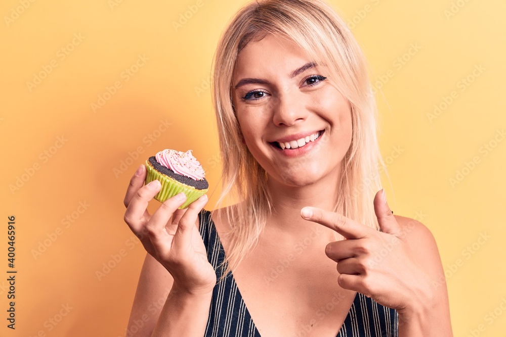 Young beautiful blonde woman eating sweet chocolate cupcake over isolated yellow background smiling happy pointing with hand and finger Young beautiful blonde woman eating sweet chocolate cupcake over isolated yellow background smiling happy pointing with hand and finger