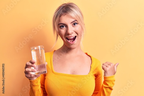 Young beautiful blonde woman drinking glass of water over isolated yellow background pointing thumb up to the side smiling happy with open mouth © Krakenimages.com