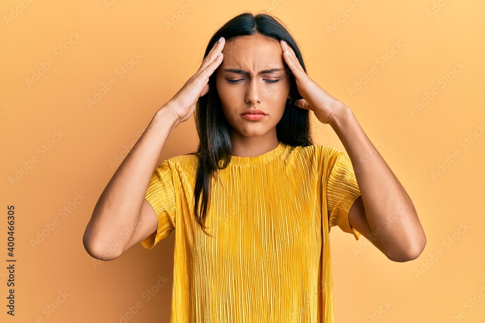 Young brunette woman wearing yellow tshirt over yellow background with hand on head, headache because stress. suffering migraine. Young brunette woman wearing yellow tshirt over yellow background with hand on head, headache because stress. suffering migraine.