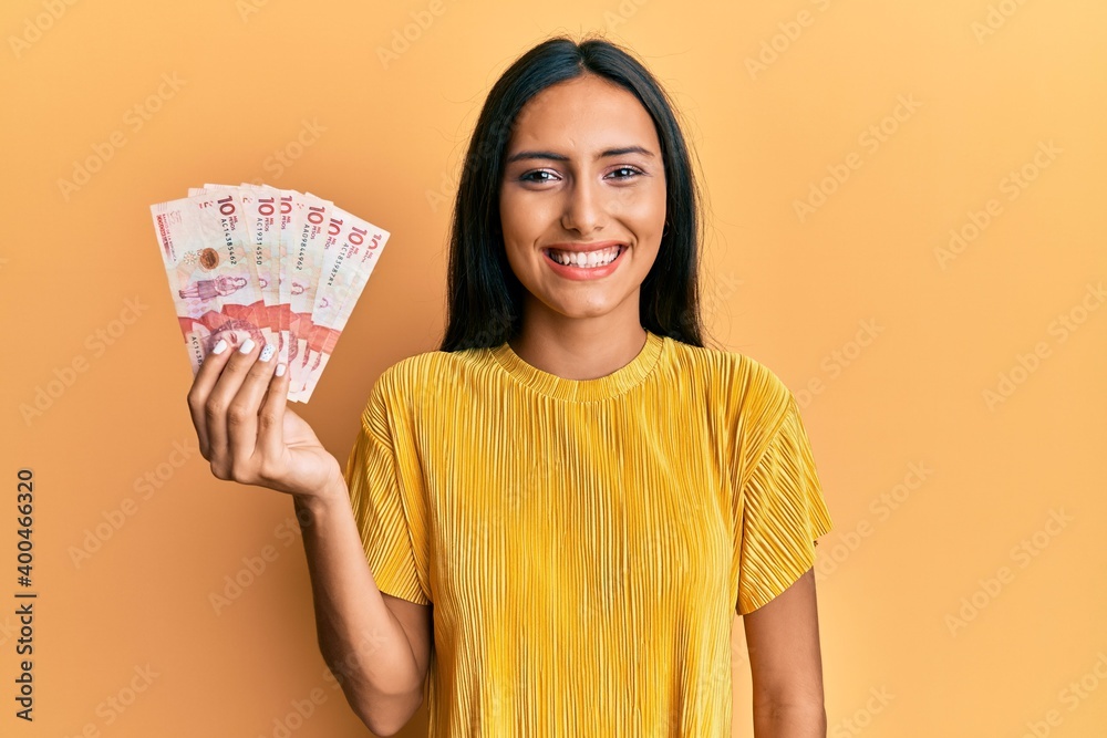Young brunette woman holding 10 colombian pesos banknotes looking positive and happy standing and smiling with a confident smile showing teeth Young brunette woman holding 10 colombian pesos banknotes looking positive and happy standing and smiling with a confident smile showing teeth