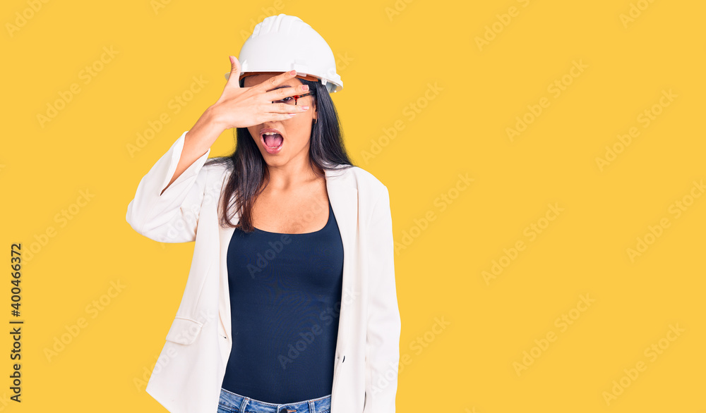 Young beautiful latin girl wearing architect hardhat and glasses peeking in shock covering face and eyes with hand, looking through fingers with embarrassed expression.