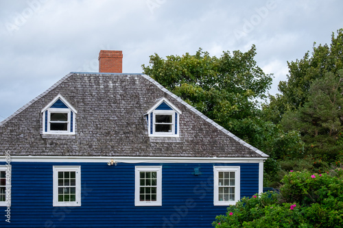 A vintage blue wooden building with white trim, double hung windows, dormers, a black shingled roof, and a brown brick chimney. The old home has multiple stories with a green tree and grey skies.