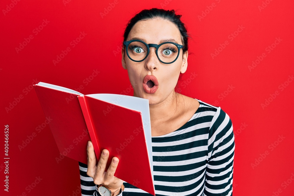 Young caucasian woman wearing glasses covering mouth with book scared and amazed with open mouth for surprise, disbelief face Young caucasian woman wearing glasses covering mouth with book scared and amazed with open mouth for surprise, disbelief face