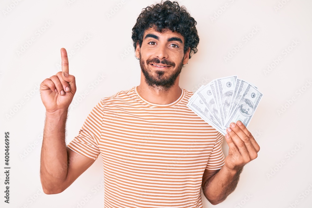 Handsome young man with curly hair and bear holding money bag with dollar symbol surprised with an idea or question pointing finger with happy face, number one Handsome young man with curly hair and bear holding money bag with dollar symbol surprised with an idea or question pointing finger with happy face, number one