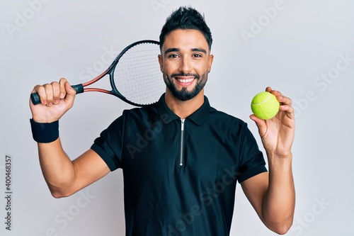 Young man with beard playing tennis holding racket and ball smiling with a happy and cool smile on face. showing teeth. © Krakenimages.com