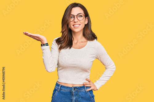 Young brunette woman wearing casual clothes and glasses smiling cheerful presenting and pointing with palm of hand looking at the camera.