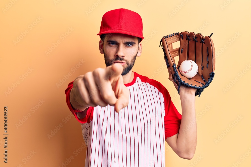 Young handsome man with beard playing baseball using ball and glove pointing with finger to the camera and to you, confident gesture looking serious