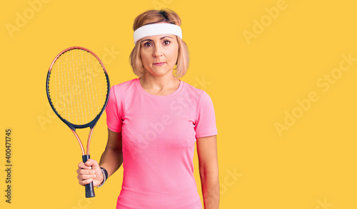 Young blonde woman playing tennis holding racket thinking attitude and sober expression looking self confident