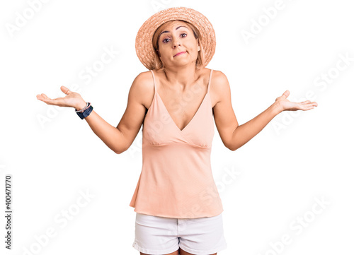 Young blonde woman wearing summer hat clueless and confused expression with arms and hands raised. doubt concept.