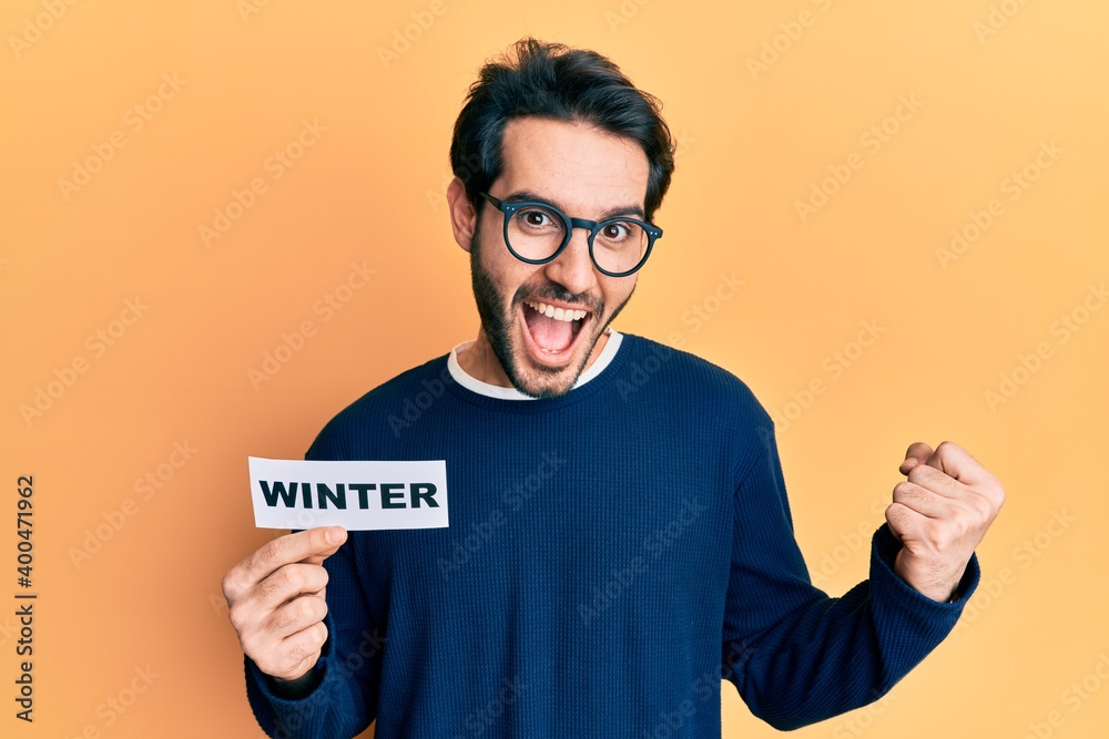 Young hispanic man wearing glasses holding winter word on paper screaming proud, celebrating victory and success very excited with raised arms Young hispanic man wearing glasses holding winter word on paper screaming proud, celebrating victory and success very excited with raised arms