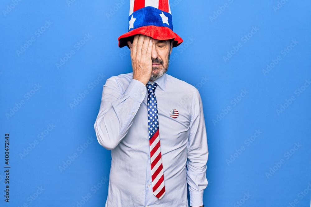Middle age handsome patriotic man wearing united states hat and tie over blue background surprised with hand on head for mistake, remember error. Forgot, bad memory concept. Middle age handsome patriotic man wearing united states hat and tie over blue background surprised with hand on head for mistake, remember error. Forgot, bad memory concept.