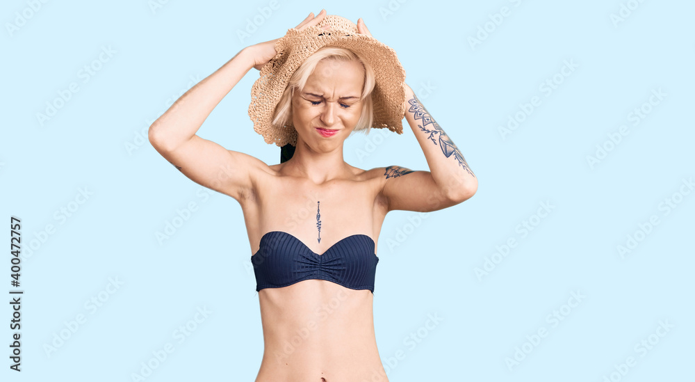 Young blonde woman with tattoo wearing bikini and summer hat suffering from headache desperate and stressed because pain and migraine. hands on head. Young blonde woman with tattoo wearing bikini and summer hat suffering from headache desperate and stressed because pain and migraine. hands on head.