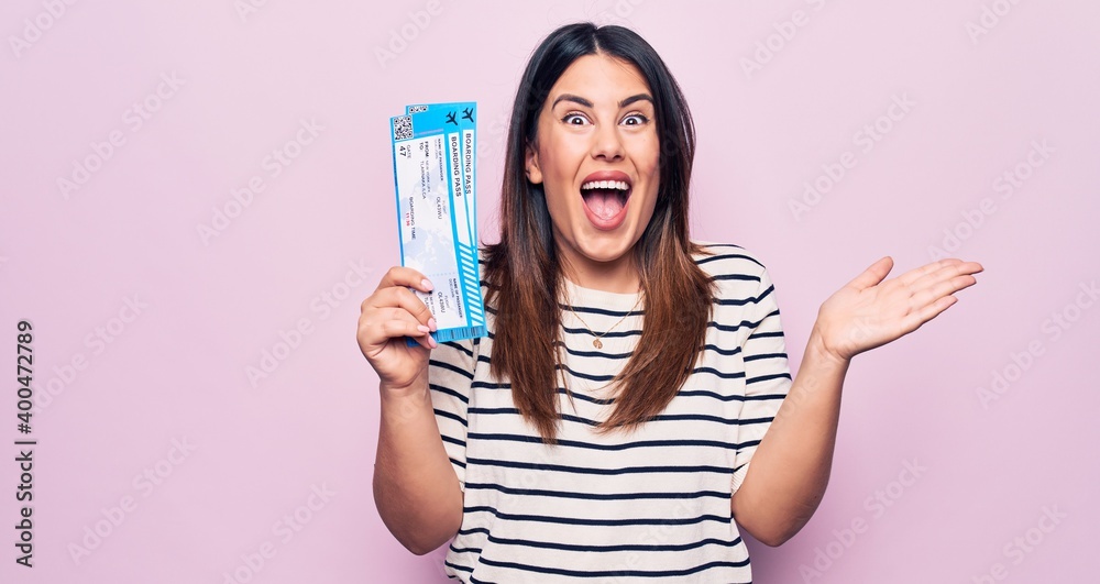 Young beautiful tourist woman holding airline boarding pass over isolated pink background celebrating achievement with happy smile and winner expression with raised hand Young beautiful tourist woman holding airline boarding pass over isolated pink background celebrating achievement with happy smile and winner expression with raised hand