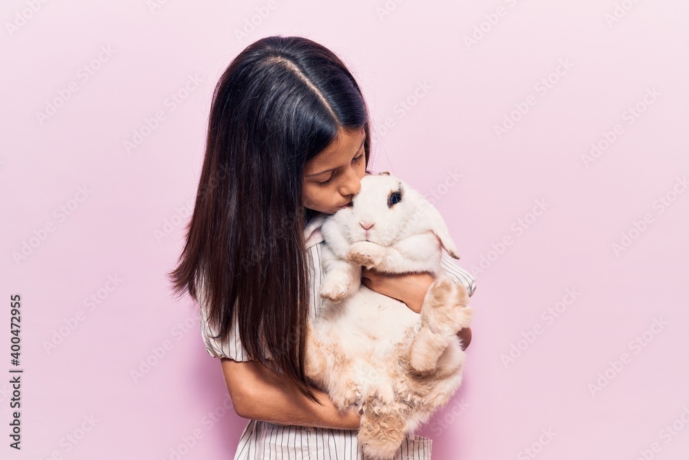 Adorable hispanic kid girl smiling happy. Holding and kissing cute rabbit standing over isolated pink background Adorable hispanic kid girl smiling happy. Holding and kissing cute rabbit standing over isolated pink background