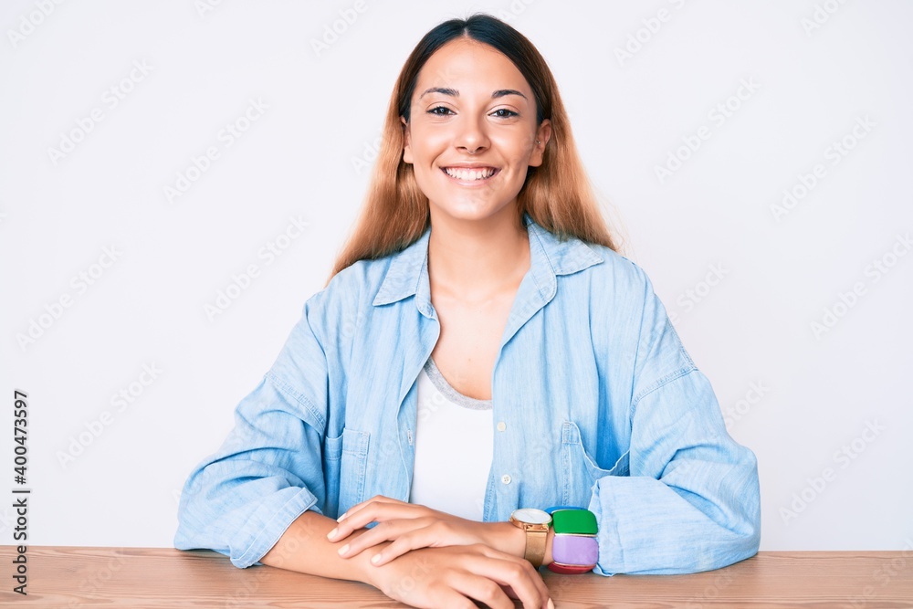 Young brunette woman sitting on the table wearing casual clothes looking positive and happy standing and smiling with a confident smile showing teeth Young brunette woman sitting on the table wearing casual clothes looking positive and happy standing and smiling with a confident smile showing teeth
