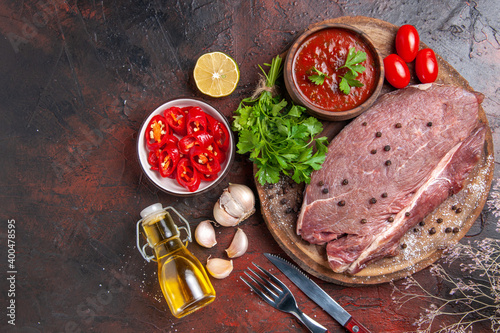 Overhead view of red meat on wooden tray and garlic green ketchup and chopped pepper fallen oil bottle on dark background