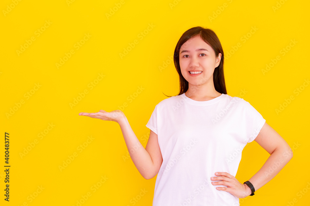 Cheerful Asian woman in white shirt standing on the yellow backgrounding studio and smiling to camera, Asian beautiful happy girl showing a hand for advertisement and artwork. Woman posing.