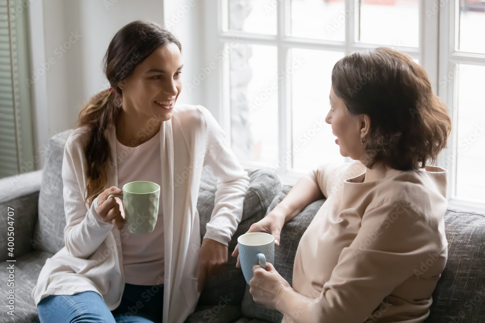 Close up happy grownup daughter and mature mother chatting, spending weekend together, sitting on cozy couch, holding cups of hot drinks, grandmother and granddaughter enjoying pleasant conversation