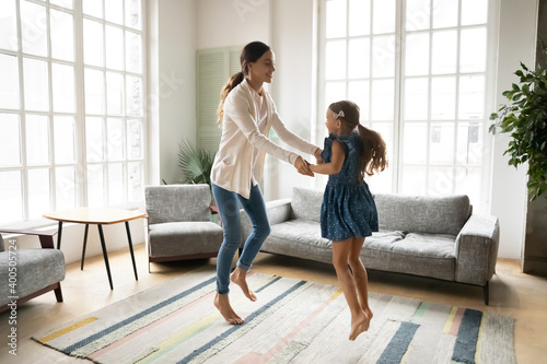 Overjoyed young mother and little daughter dancing, moving to music in living room, family enjoying leisure activity, happy mum and adorable girl child holding hands, jumping, having fun together