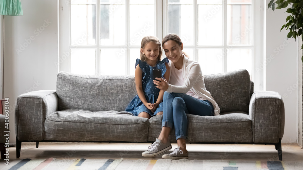 Happy young mother with little daughter using smartphone together, smiling girl child and mum looking at screen, browsing apps, shopping or chatting online, having fun with phone, making video call