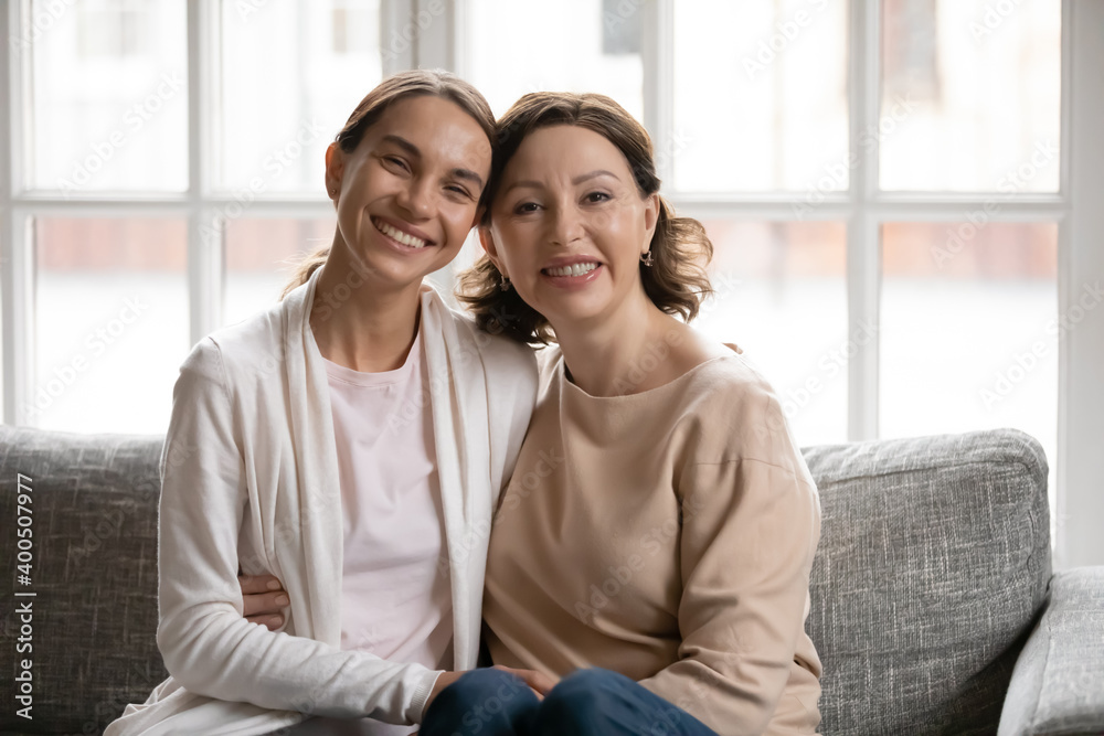 Head shot portrait smiling middle aged mother and grownup daughter hugging, sitting on cozy couch at home, happy young woman with mature mum posing for family photo together, looking at camera Head shot portrait smiling middle aged mother and grownup daughter hugging, sitting on cozy couch at home, happy young woman with mature mum posing for family photo together, looking at camera