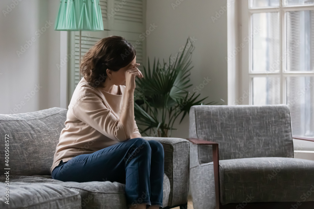 Fototapeta premium Depressed pensive middle aged woman lost in thoughts, sitting on couch at home alone, looking in distance, upset mature female thinking about problems, touching forehead, suffering from headache