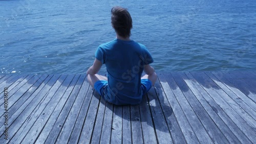 Young sporty man sitting in meditative pose on wooden pier looking at waves at daytime. Man in blue in windy weather. High quality 4k footage