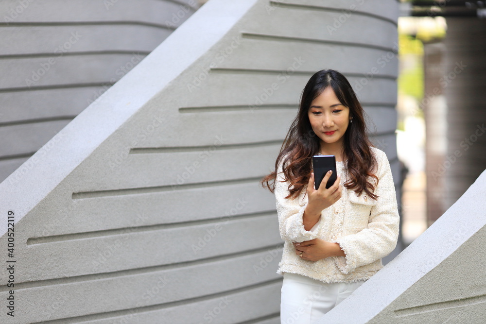 Fototapeta premium Asian lady using mobile phone while walking along the modern building in urban downtown area for communication and technology concept