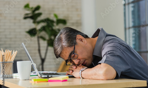 Asian businessman tired overworked he sleeping over a laptop computer on the desk. senior man with eyeglasses lying asleep on table at his working place
