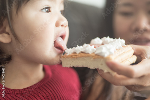 A toddler sitting on a sofa sticks her tongue out to lick a piece of sponge cake with cream held by her mother in a house in Edinburgh, Scotland, United Kingdom