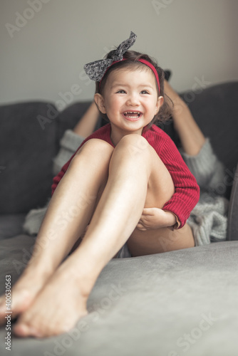 A toddler wearing a red top and a hair band with a hair laughs and plays on top on her mother   s legs while sitting on a sofa in a house in Edinburgh  Scotland  United Kingdom
