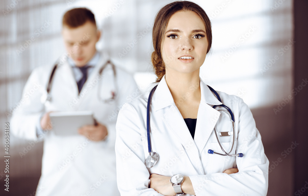 Portrait of a professional friendly woman doctor with a stethoscope, standing with crossed arms in a clinic. Young doctors at work in a hospital. Medicine and healthcare concept Portrait of a professional friendly woman doctor with a stethoscope, standing with crossed arms in a clinic. Young doctors at work in a hospital. Medicine and healthcare concept