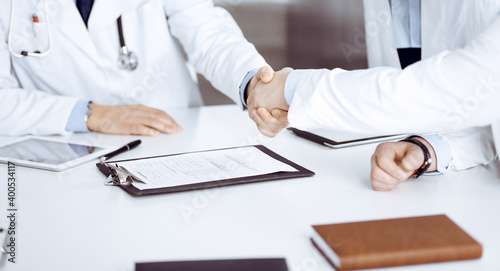 Unknown doctors are shaking their hands finishing discussion about patient's diagnosis, sitting at the desk and using a clipboard, close-up. Medical help, insurance in health care, medicine concept