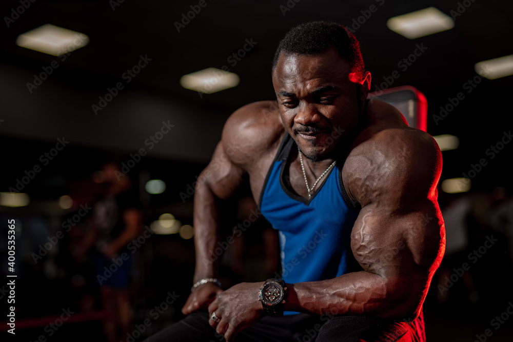 African american bodybuilder man poses to the camera in gym. Big muscles and strong hands. Blurred background. African american bodybuilder man poses to the camera in gym. Big muscles and strong hands. Blurred background.