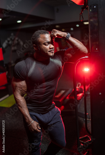 Fitness trainer poses to the camera in modern gym with hand near face. Perfect body of professonal bodybuilder. photo