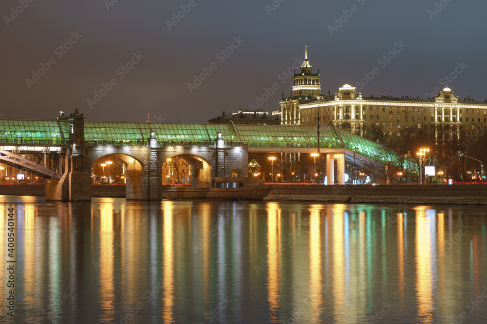 Moscow bright illuminated cityscape. Andreevsky (Pushkinsky) pedestrian bridge, wide Moskva river, Stalinist architecture. Dark winter night sky during coronavirus pandemic