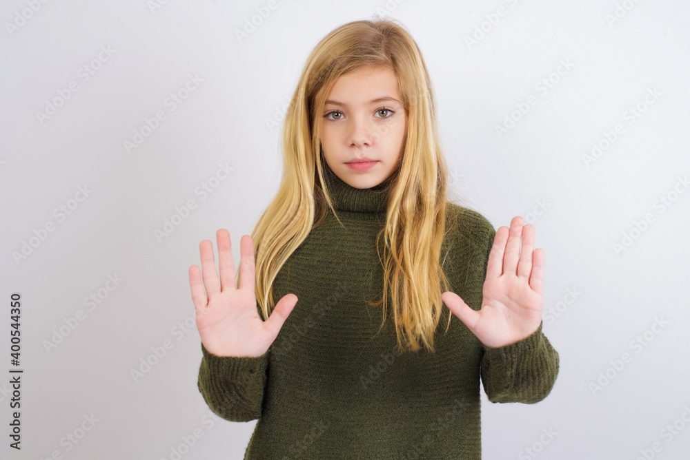 Serious Caucasian kid girl wearing green knitted sweater against white wall pulls palms towards camera, makes stop gesture, asks to control your emotions and not be nervous Serious Caucasian kid girl wearing green knitted sweater against white wall pulls palms towards camera, makes stop gesture, asks to control your emotions and not be nervous