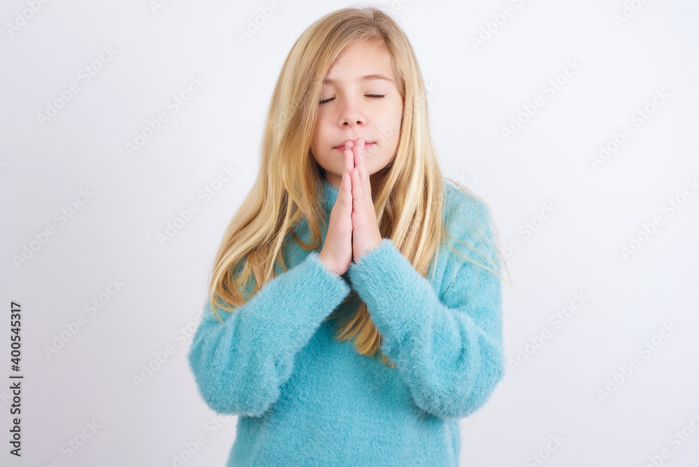 Indoor closeup of Cute Caucasian kid girl wearing blue knitted sweater against white wall practicing yoga and meditation, holding palms together in namaste, looking calm, relaxed and peaceful. Indoor closeup of Cute Caucasian kid girl wearing blue knitted sweater against white wall practicing yoga and meditation, holding palms together in namaste, looking calm, relaxed and peaceful.