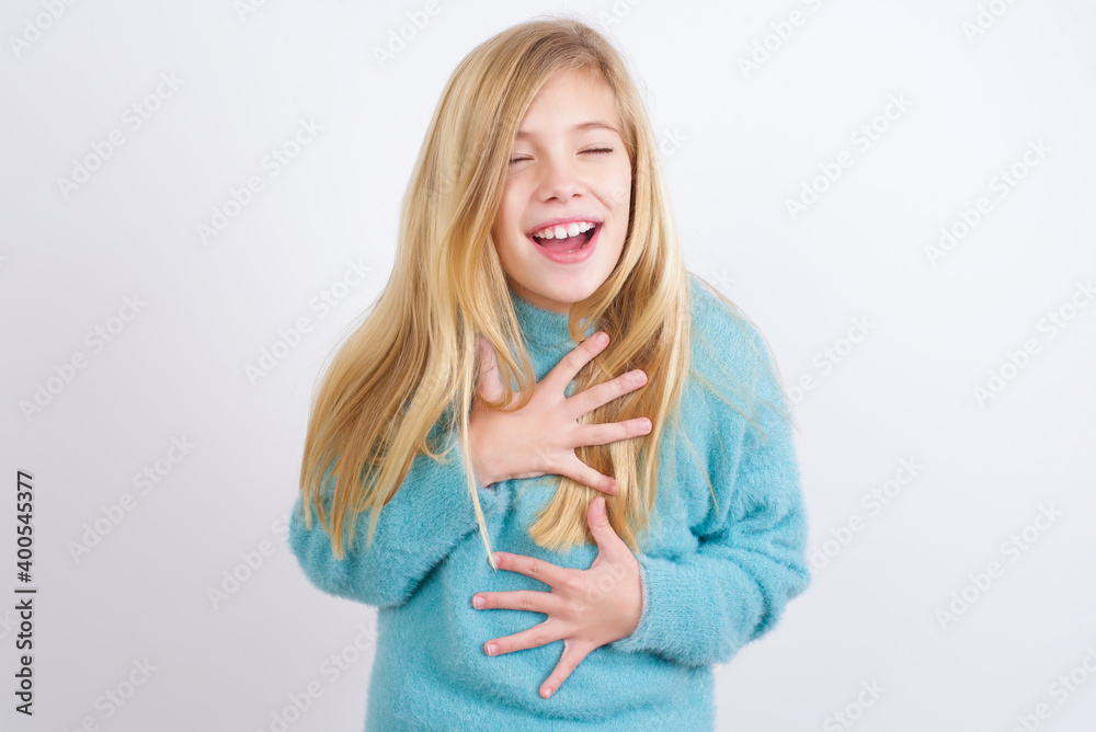 Cute Caucasian kid girl wearing blue knitted sweater against white wall keeps hands crossed, laughs out loud at good joke, wears casual clothes. Cute Caucasian kid girl wearing blue knitted sweater against white wall keeps hands crossed, laughs out loud at good joke, wears casual clothes.