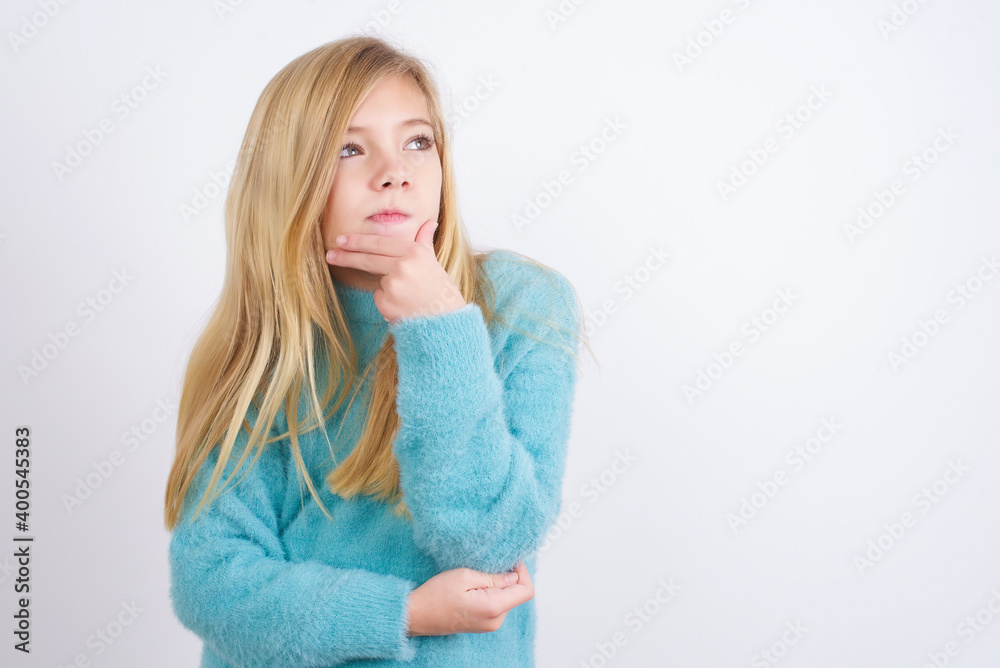 Shot of contemplative thoughtful Cute Caucasian kid girl wearing blue knitted sweater against white wall keeps hand under chin, looks thoughtfully upwards. Shot of contemplative thoughtful Cute Caucasian kid girl wearing blue knitted sweater against white wall keeps hand under chin, looks thoughtfully upwards.