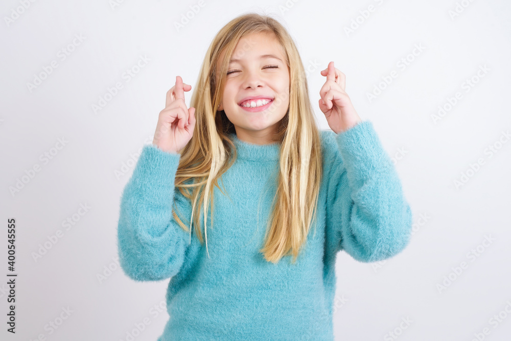 Joyful Cute Caucasian kid girl wearing blue knitted sweater against white wall clenches teeth, raises fingers crossed, makes desirable wish, waits for good news, I have to win. Joyful Cute Caucasian kid girl wearing blue knitted sweater against white wall clenches teeth, raises fingers crossed, makes desirable wish, waits for good news, I have to win.