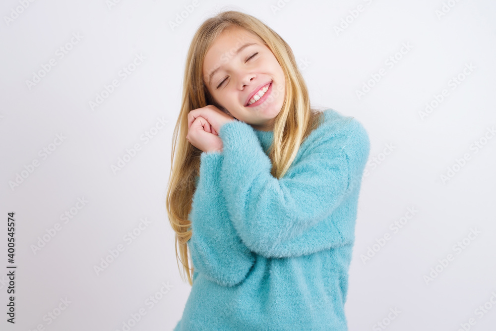 Dreamy Cute Caucasian kid girl wearing blue knitted sweater against white wall with pleasant expression, closes eyes, keeps hands crossed near face, thinks about something pleasant
