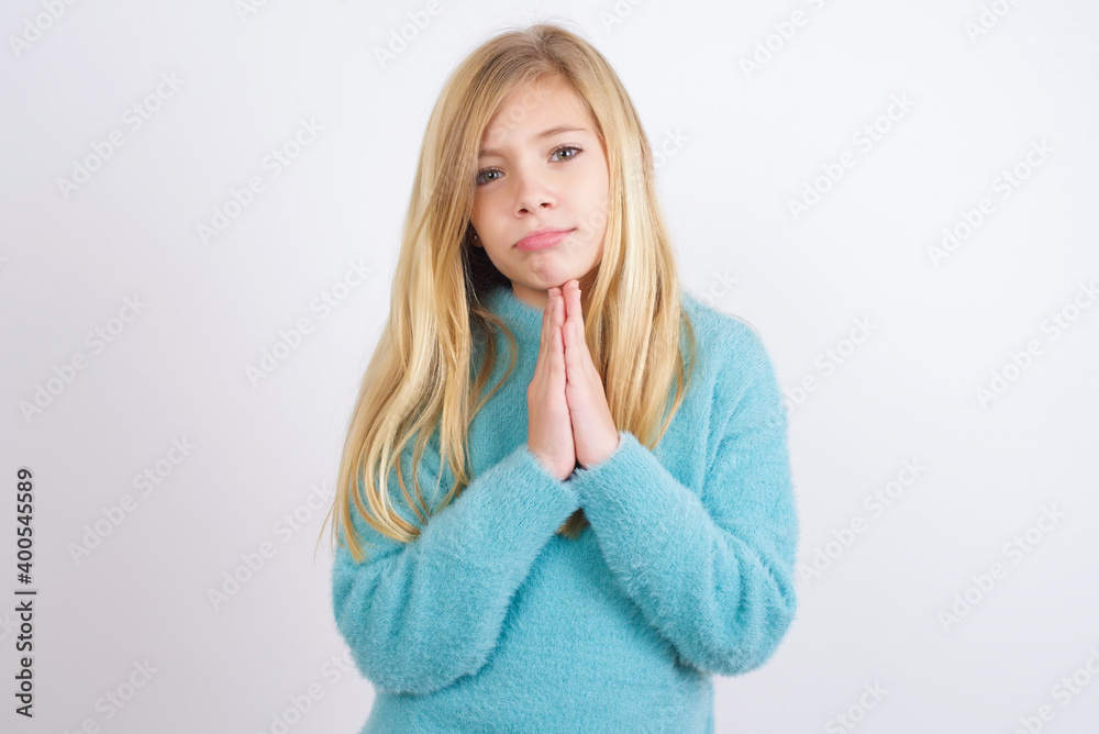 Cute Caucasian kid girl wearing blue knitted sweater against white wall keeps palms pressed together in front of her having regretful look, asking for forgiveness. Forgive me please. Cute Caucasian kid girl wearing blue knitted sweater against white wall keeps palms pressed together in front of her having regretful look, asking for forgiveness. Forgive me please.