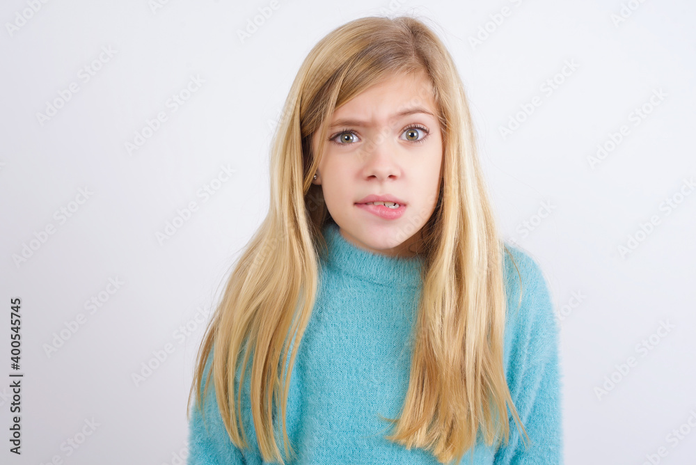Cute Caucasian kid girl wearing blue knitted sweater against white wall being nervous and scared biting lips looking camera with impatient expression, pensive. Cute Caucasian kid girl wearing blue knitted sweater against white wall being nervous and scared biting lips looking camera with impatient expression, pensive.