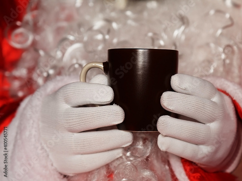Santa Claus holding hands in a dark brown mug of tea in front of a white beard