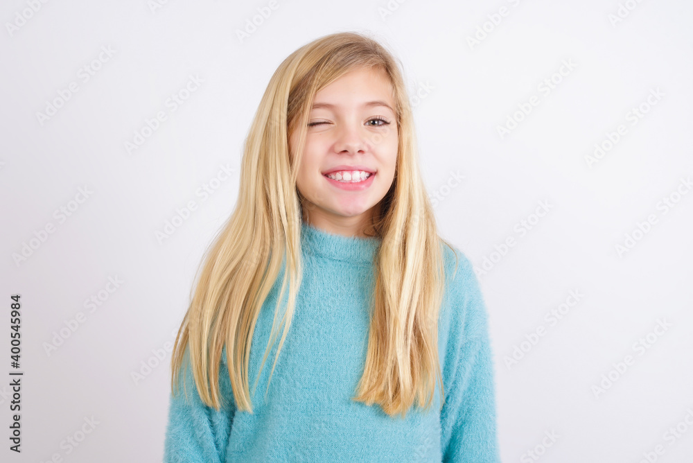 Cute Caucasian kid girl wearing blue knitted sweater against white wall blinking eyes with pleasure having happy expression. Facial expressions and people emotions concept. Cute Caucasian kid girl wearing blue knitted sweater against white wall blinking eyes with pleasure having happy expression. Facial expressions and people emotions concept.