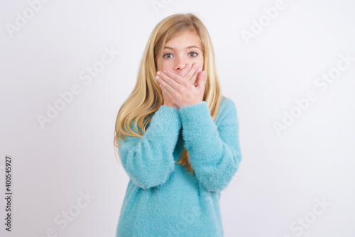 Upset Cute Caucasian kid girl wearing blue knitted sweater against white wall, covering her mouth with both palms to prevent screaming sound, after seeing or hearing something bad.