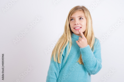 Cute Caucasian kid girl wearing blue knitted sweater against white wall with thoughtful expression, looks to the camera, keeps hand near face, bitting a finger thinks about something pleasant.